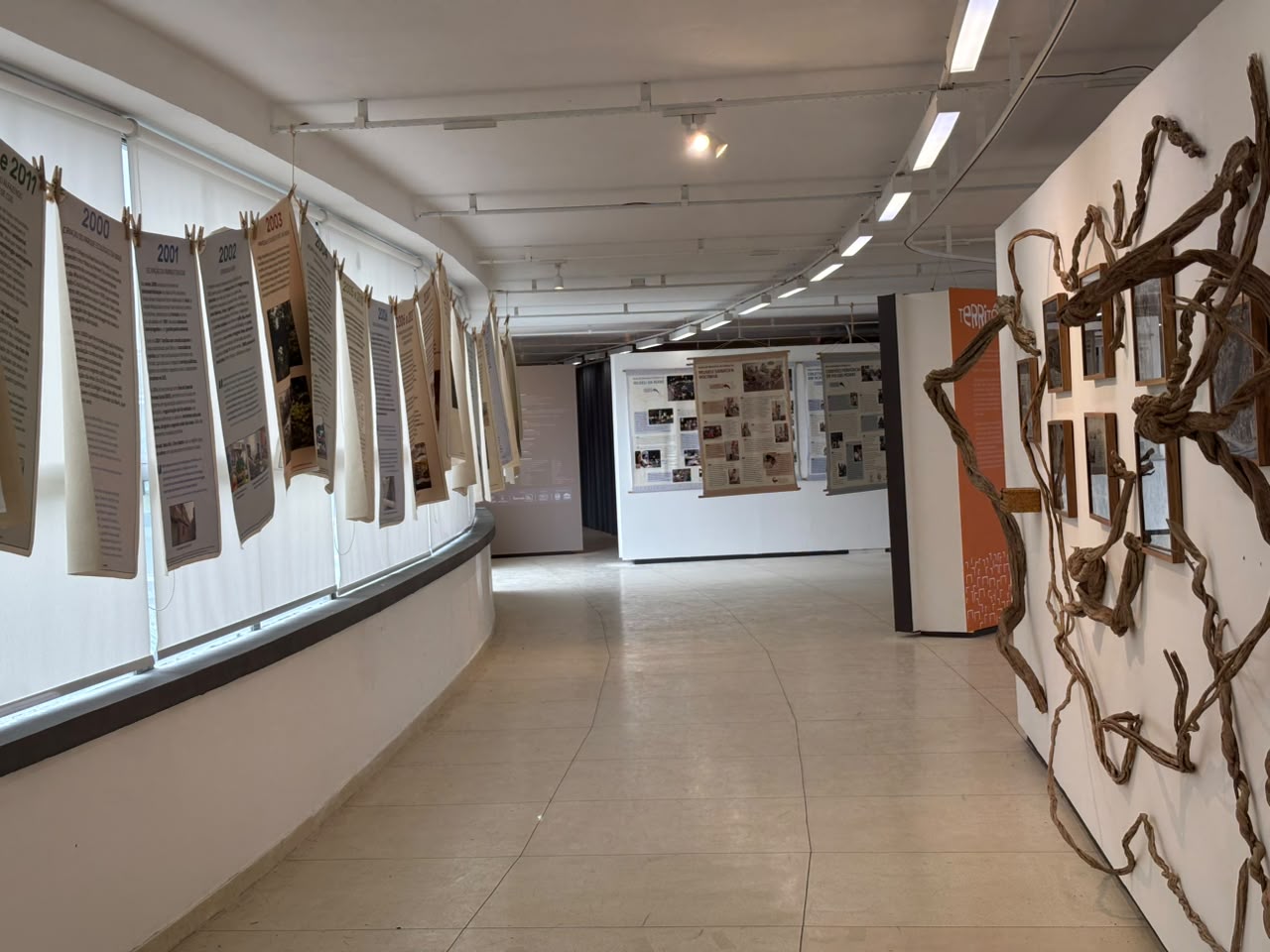 Corredor expositivo com linha do tempo, painéis suspensos e instalação artística na exposição Memória Climática das Favelas.